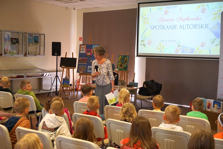 Jedna z zaproszonych kobiet w jednej ręce trzyma ksiażkę w drugiej mikrofon, przechadza się między rzędami krzeseł na których siedzą dzieci.