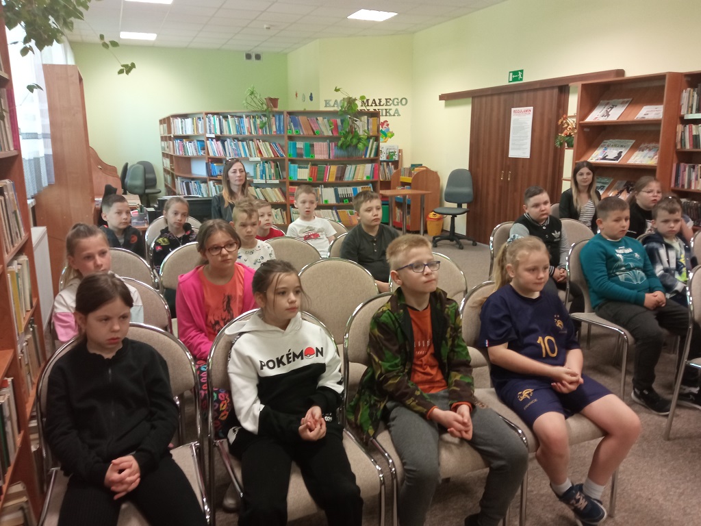 Dzieci i nauczycielki obecne na spotkaniu siedzą na krzesłach i słuchają zaproszonych gościń