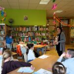 dzieci siedzące przy stołach patrzą w stronę bibliotekarki która pokazuje im okładkę książki.