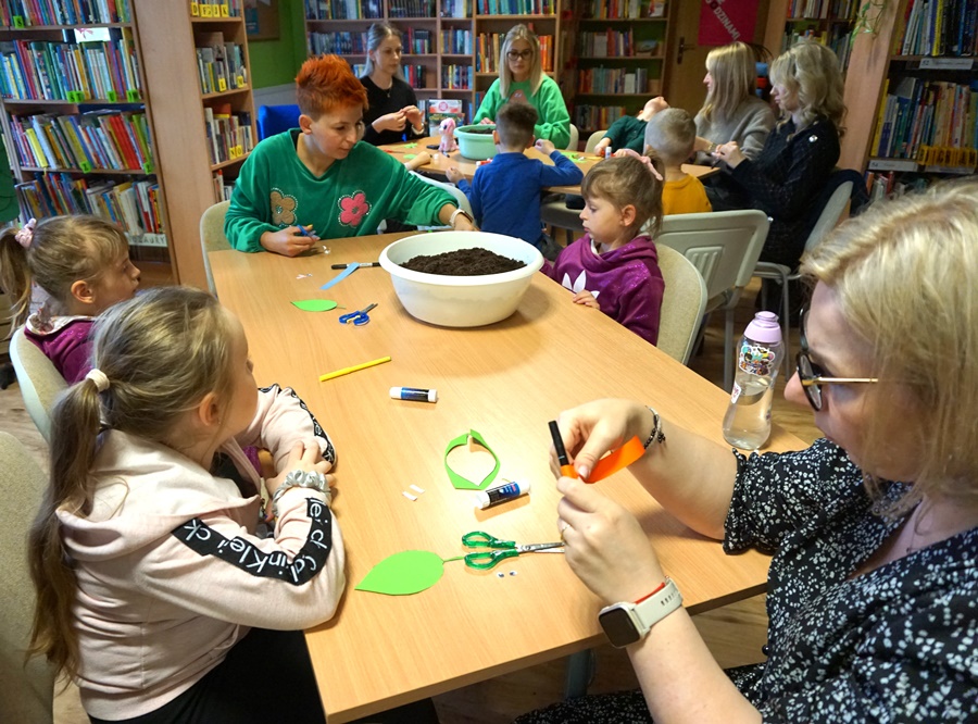 dzieci z rodzicami siedzą przy stołach i sklejają elementy z papieru.