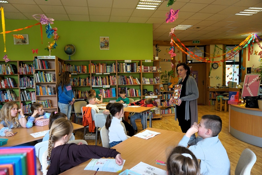 dzieci siedzące przy stołach patrzą w stronę bibliotekarki która pokazuje im okładkę książki.