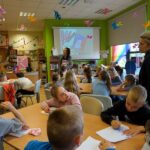 Dzieci siedzą przy stolikach, nauczycielka obserwuje grupę, bibliotekarka pokazuje coś dziciom