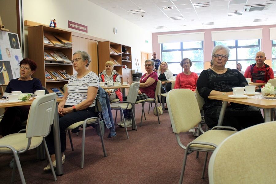 Część publiczności, ludzie siedzący przy stolikach, na których stoją ciastka i filiżanki na kawę lub herbatę.