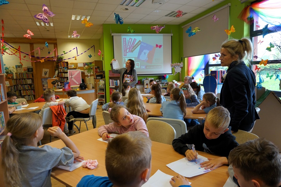 Dzieci siedzą przy stolikach, nauczycielka obserwuje grupę, bibliotekarka pokazuje coś dziciom