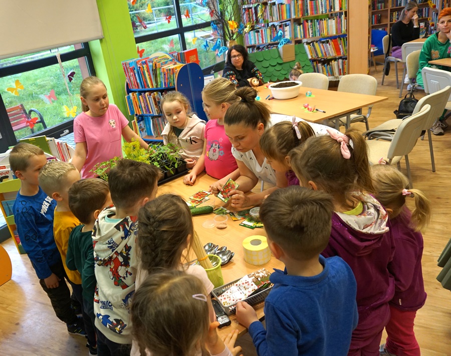 dzieci zebrane wokół stołu przy którym bibliotekarka pokazuje i tłumaczy to jakie nasiona znajdują sięw konkretnej paczuszce.