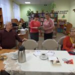 5 osób, 4 kobiety i mężczyzna, dwie kobiety stoją, pozostali siedzą przy stole, jedn ze stojących kobiet trzyma w ręku książki, druga przygląda się tym książkom