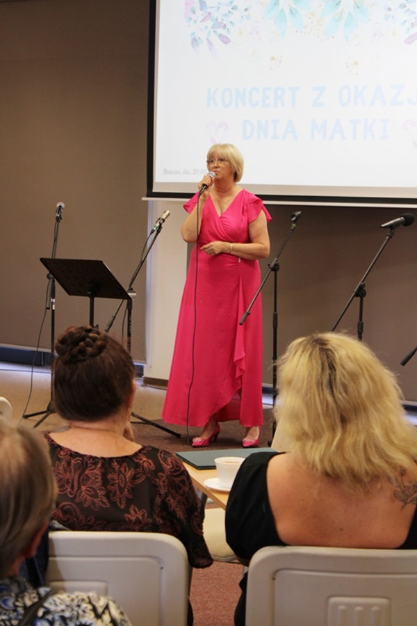 Nell Polgert-Martwijczuk (wysoka blondynka z krótkimi włosami ubrana w różową suknię) w czasie występu stoi przy mikrofonie i śpiewa przed cebraną publicznością