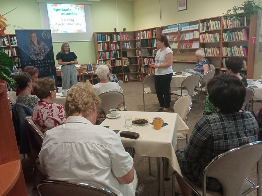 Widownia zebrana na spotkaniu siedzi przy stoikach, kierowniczka filii w Piechcinie stoi przy stoliku i wita zebrane osoby, zaproszona autorka stoi przed zebraną publicznością