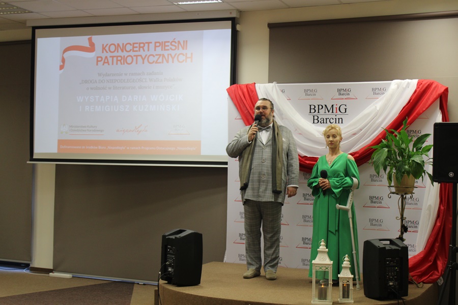 Duet wokalistów stojący na scenie, pan Remigiusz mówi coś do mikrofonu