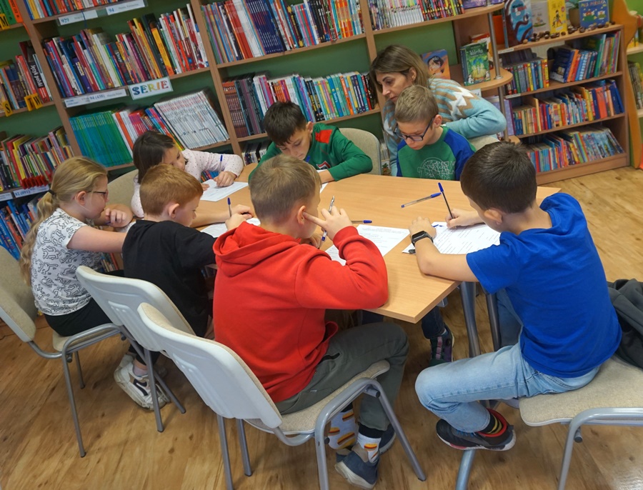 dzieci z opiekunami siedzą przy stolikach i piszą coś na kartkach papieru