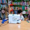 dwie dziewczynki razem z bibliotekarką siedzą przy stole na którym stoi maszyna do szycia, widoczne osoby wycinają coś z materiału