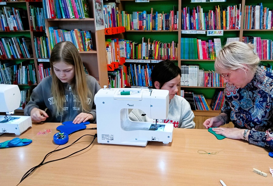 dwie dziewczynki razem z bibliotekarką siedzą przy stole na którym stoi maszyna do szycia, widoczne osoby wycinają coś z materiału