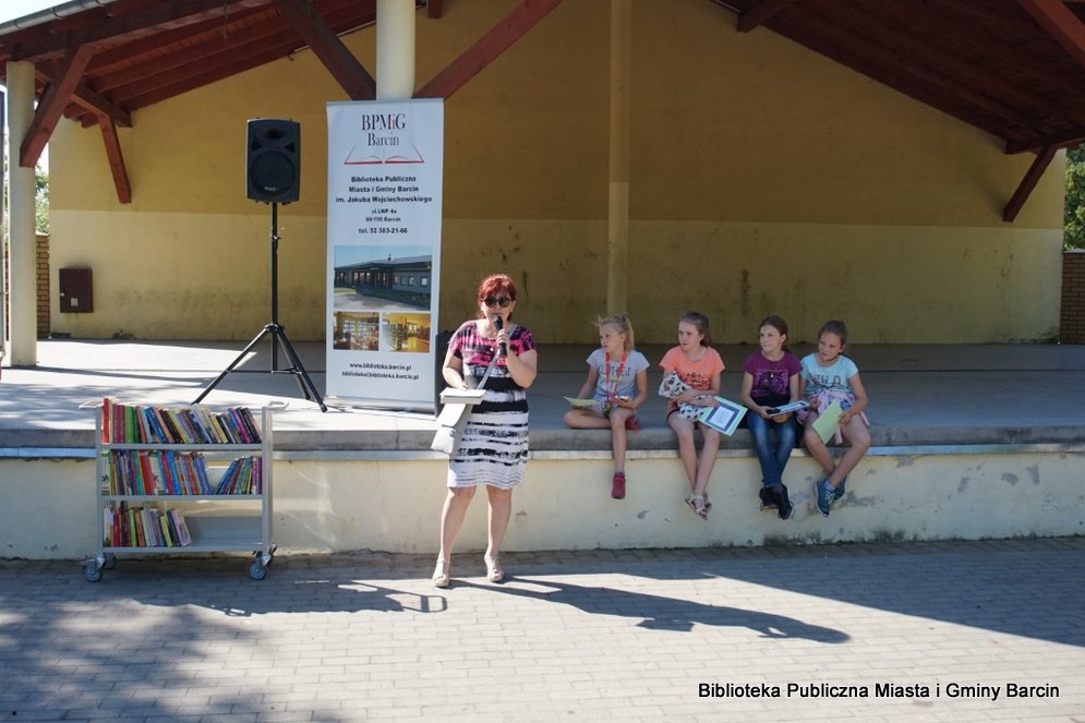 Kierownik filii w Piechcinie stoi z mikrofonem w ręce i rozpoczyna spotkanie za nią na brzegu sceny siedzą 4 dziewczynki pomagające w wydarzeniu