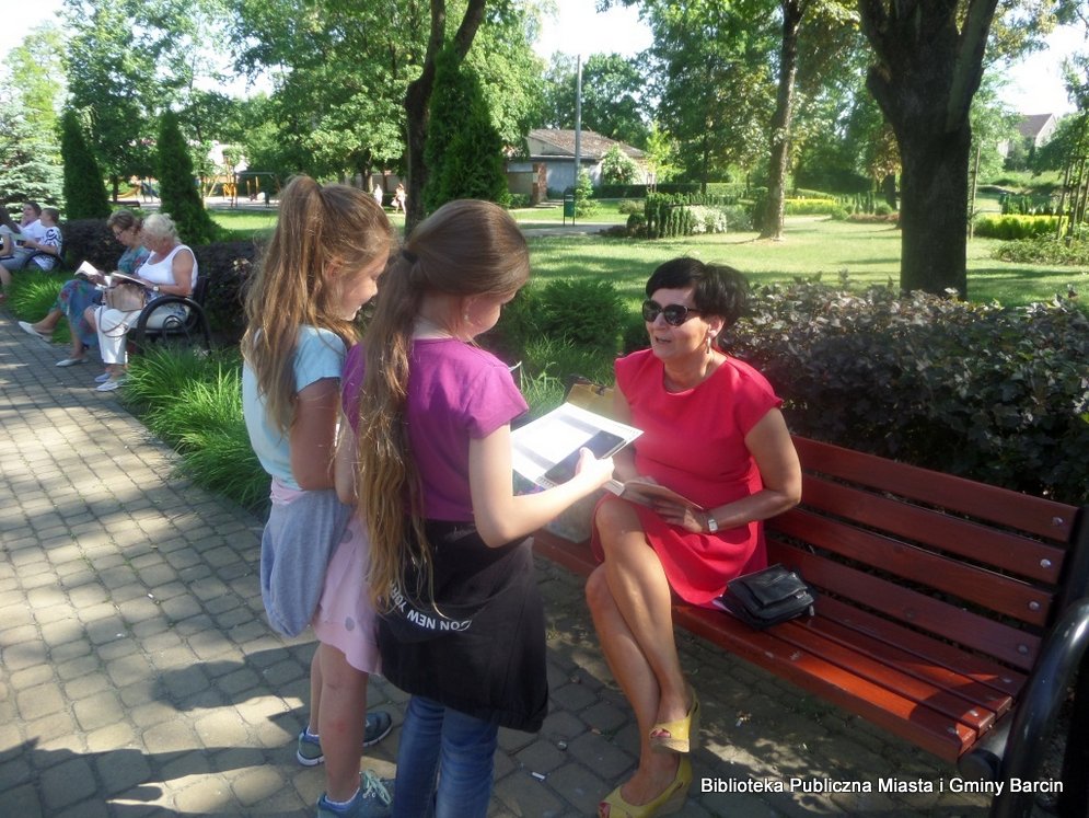 2 dziewczynki stoją przed kobietą siedzącą na ławce i przeprowadzają z nią ankietę