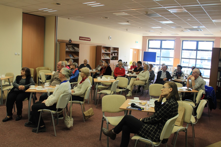 Publiczność zebrana w bibliotece siedzi przy stolikach, popija kawę lub herbatę i w skupieniu słucha twórczości zaproszonego gościa.