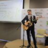po lewej zaproszony gość stoi przy mikrofonie z gitarą w rękach, po lewej widoczny jest ekran z plakatem promującym spotkanie