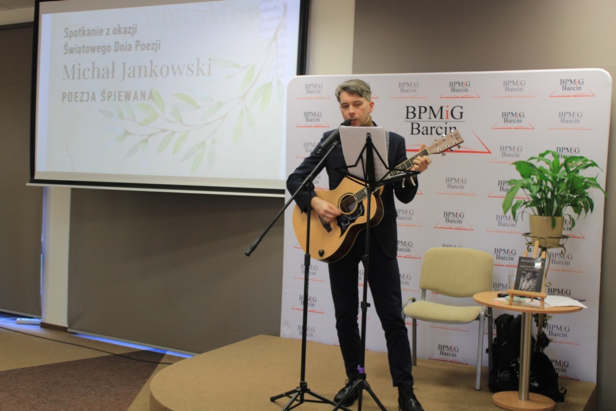po lewej zaproszony gość stoi przy mikrofonie z gitarą w rękach, po lewej widoczny jest ekran z plakatem promującym spotkanie
