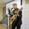 Zaproszony gość z gitarą w rękach stoi przy mikrofonie i śpiewa swój wiersz.
