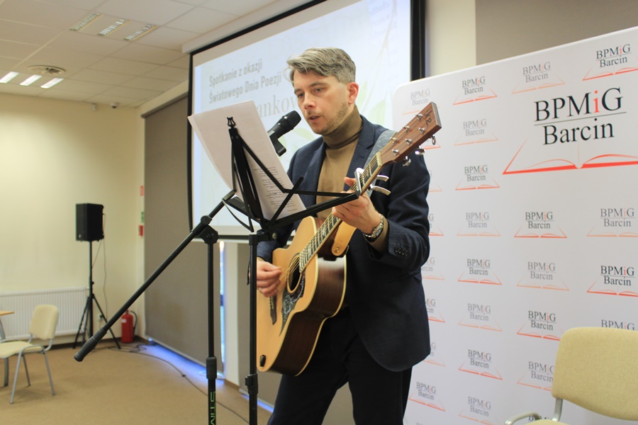 Zaproszony gość z gitarą w rękach stoi przy mikrofonie i śpiewa swój wiersz.