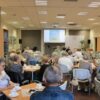 Widownia zebrana w bibliotece, ludzie siedzący na krzesłach przy kwadratowych stolikach