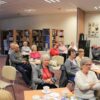 Widownia zebrana na spotkaniu, ludzie siedzący przy kwadratowych stolikach z kawą i herbatą