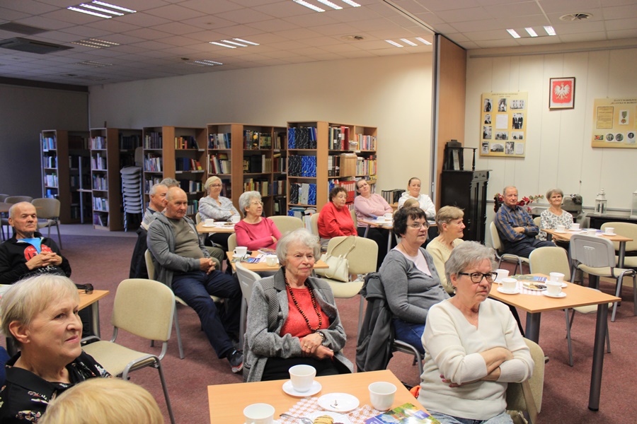 Widownia zebrana na spotkaniu, ludzie siedzący przy kwadratowych stolikach z kawą i herbatą