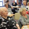Widownia zebrana w bibliotece, ludzie siedzący na krzesłach przy kwadratowych stolikach