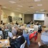 Widownia zebrana w bibliotece, ludzie siedzący na krzesłach przy kwadratowych stolikach