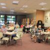 Widownia zebrana na spotkaniu, ludzie siedzący przy kwadratowych stolikach z kawą i herbatą