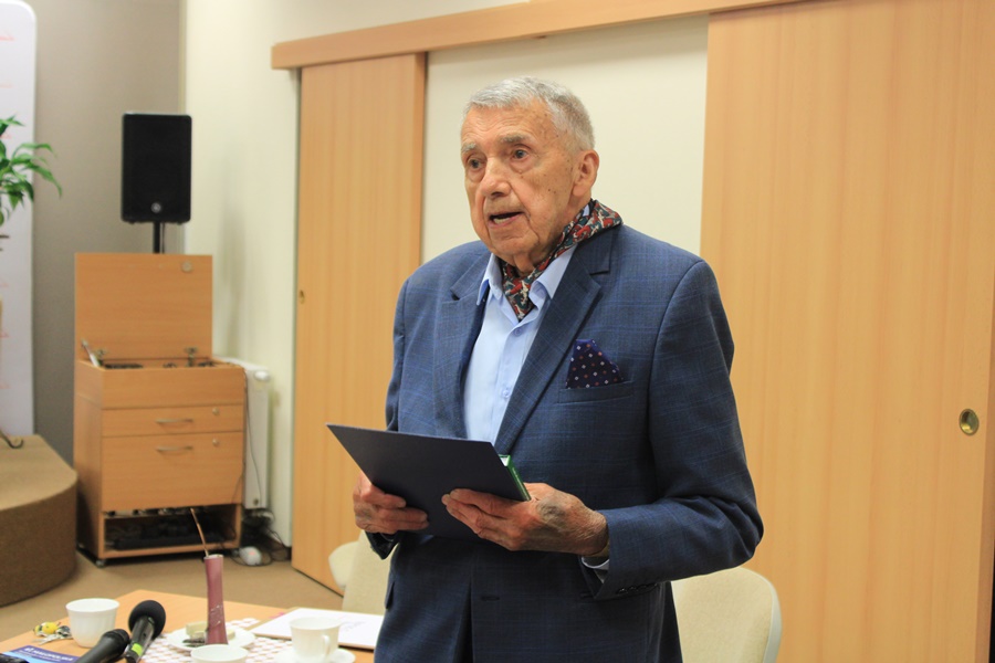 Pan Kaszyński, starszy mężczyzna w granatowym garniturze i niebieskiej koszuli trzyma w rękach otrzymaną w prezencie książkę