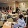 Widownia zebrana na spotkaniu, ludzie siedzący przy kwadratowych stolikach z kawą i herbatą