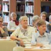 Widownia zebrana w bibliotece, ludzie siedzący na krzesłach przy kwadratowych stolikach