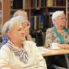 Widownia zebrana w bibliotece, ludzie siedzący na krzesłach przy kwadratowych stolikach