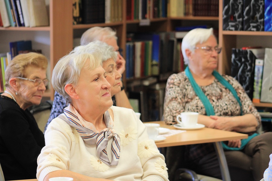 Widownia zebrana w bibliotece, ludzie siedzący na krzesłach przy kwadratowych stolikach
