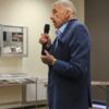 Pan Kaszyński, starszy mężczyzna w granatowym garniturze i niebieskiej koszuli trzyma w ręce mikrofon