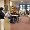 po lewej dyrektorka biblioteki z mikrofonem w ręce, w centralnej części uczestnicy siedzący w drużynach przy stolikach za nimi widzowie