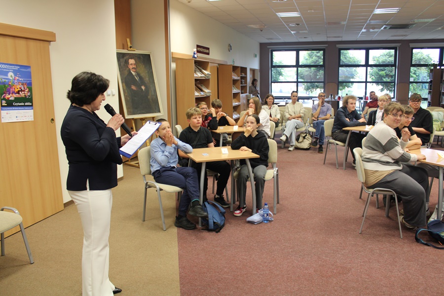 po lewej dyrektorka biblioteki z mikrofonem w ręce, w centralnej części uczestnicy siedzący w drużynach przy stolikach za nimi widzowie