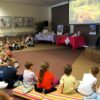 dzieci siedzą na materacach, oglądają prezentacje i słuchają opowieści zaproszonego gościa