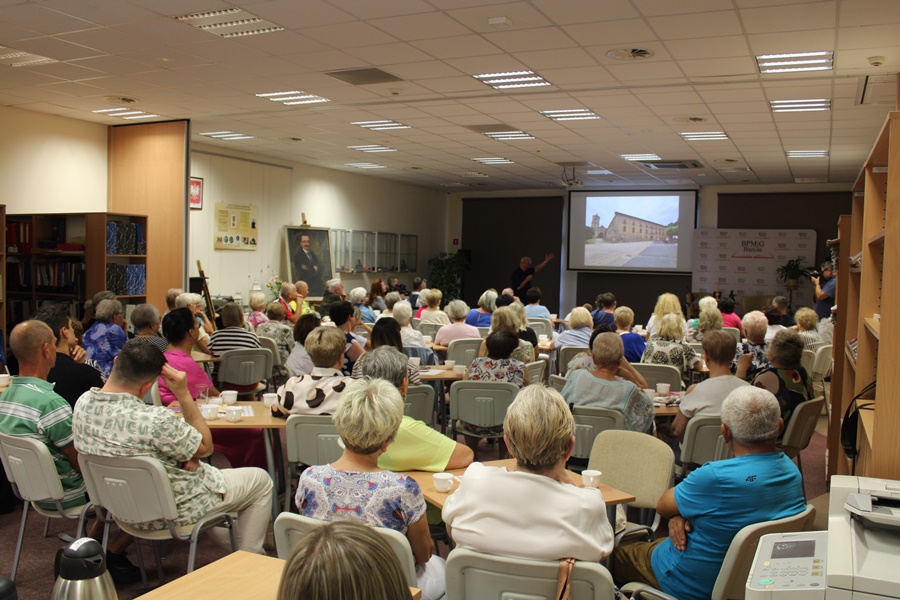 widoczna na zdjęciu pełna sala, ludzie siedzą przy stolikach, niektórzy tylko na krzesłach, wszyscy z uwagąsłuchają występującej przed nimi osoby
