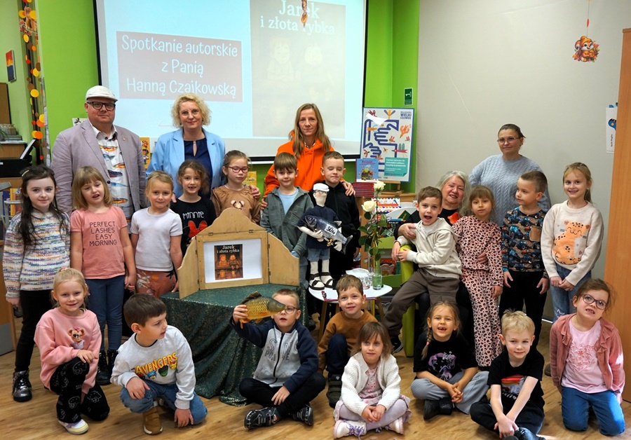 Grupowe zdjęcie uczestników zajęć, dorośli i dzieci stoją w trzech rzędach, pomiędzy nimi na stoliku stoi drewniany teatrzyk z kolorowymi obrazkami, jedno z dzieci trzyma zabawkę w kształcie ryby, inne kukiełkę przedstawiającą mężczyznę z wędką i rybą