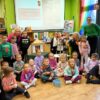 Grupowe zdjęcie grupy uczestniczącej w zajęciach, część dzieci siedzi na podłodze w pozycji po turecku inne stoją, razem z nimi stoi bibliotekarka i opiekunka grupy, zaproszona na spotkanie autorka siedzi między dziećmi na krześle