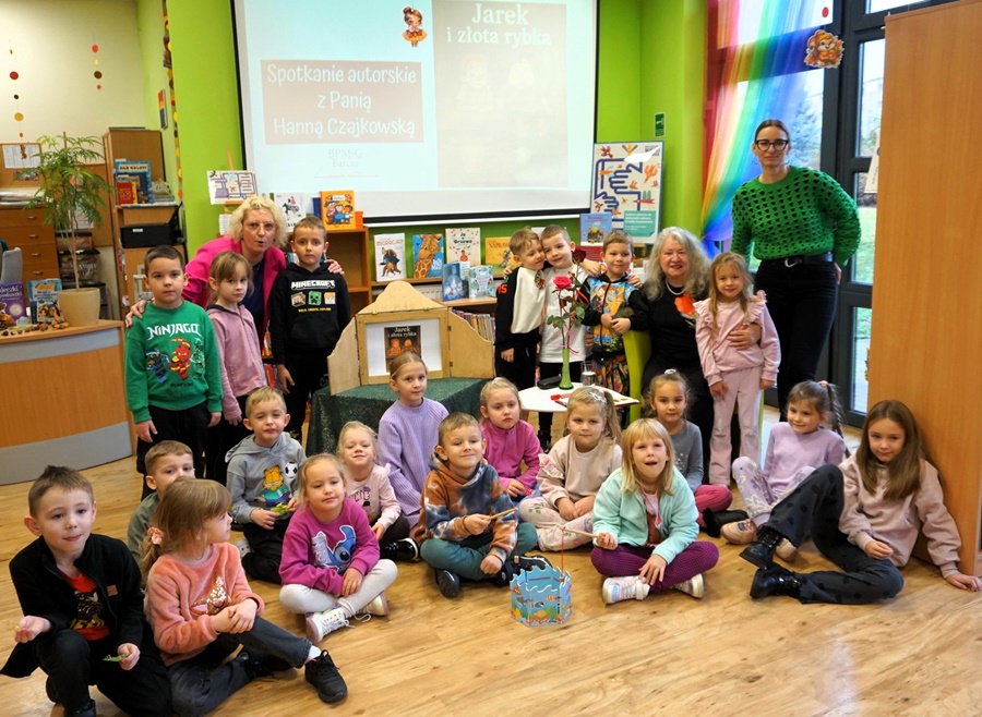 Grupowe zdjęcie grupy uczestniczącej w zajęciach, część dzieci siedzi na podłodze w pozycji po turecku inne stoją, razem z nimi stoi bibliotekarka i opiekunka grupy, zaproszona na spotkanie autorka siedzi między dziećmi na krześle