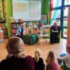 Zaproszona autorka siedzi na fotelu przed dziećmi, obok niej na stoliku stoi drewniany teatrzyk przy którym kuca bibliotekarka prowadząca zajęcia