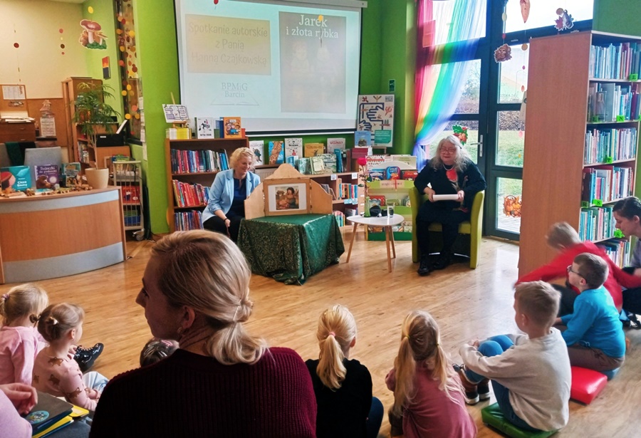 Zaproszona autorka siedzi na fotelu przed dziećmi, obok niej na stoliku stoi drewniany teatrzyk przy którym kuca bibliotekarka prowadząca zajęcia
