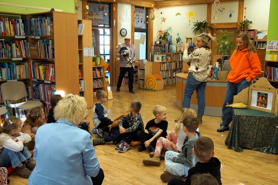 dzieci siedzą w okręgu na pufkach, w środku kuca bibliotekarka, za dziećmi stoją opiekunki, do sali wchodzi zaproszony gość z kukiełką na rękach