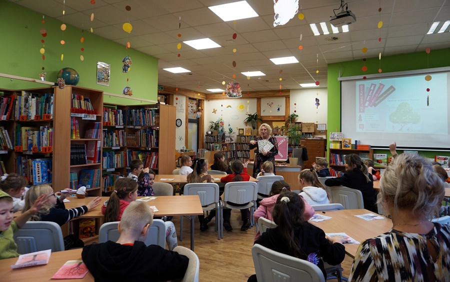 Bibliotekarka stoi przed zebraną grupa dzieci i pokazuje im ilustracje