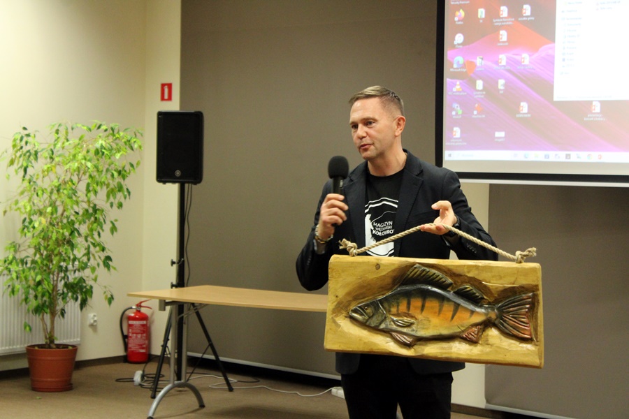 Pan Adam tański stoi pzred publicznością w jednej ręce trzyma mikrofon a w drugiej płaskorzeźbę ryby