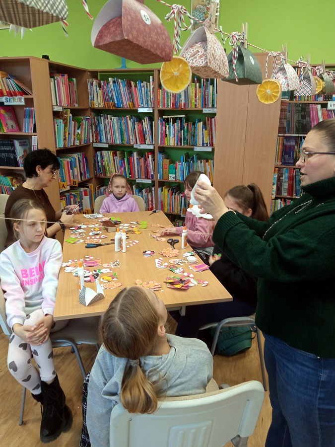 Część dzieci siedzi przy stole i wykonuje ozdoby a bibliotekarka mocuje ozdoby na sznurku