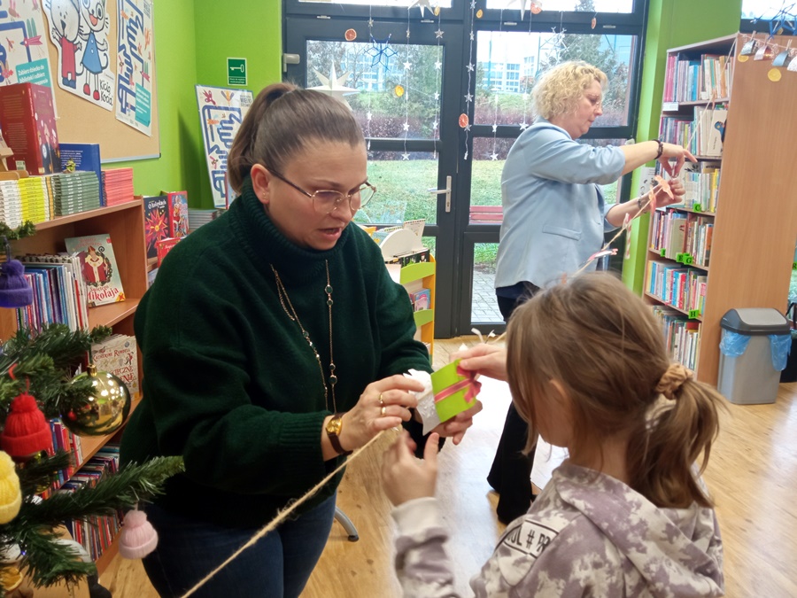 Bibliotekarka pomaga i pokazuje dziewczynce jak należy mocować wykonaną ozdobę na sznurku