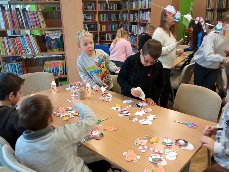 Dzieci stoją lub siedzą przy stole i sklejają ozdoby świąteczne